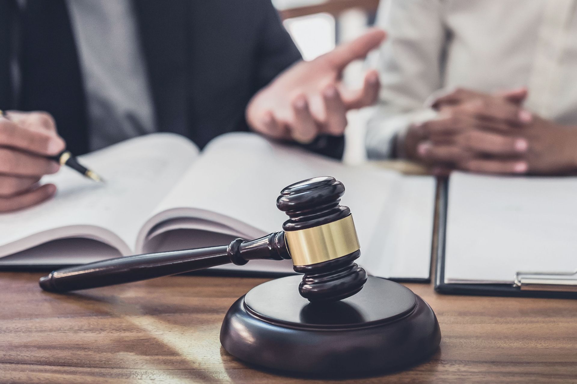 Gavel on desk with open book; two people in business attire, one pointing and discussing, other holding notepad.