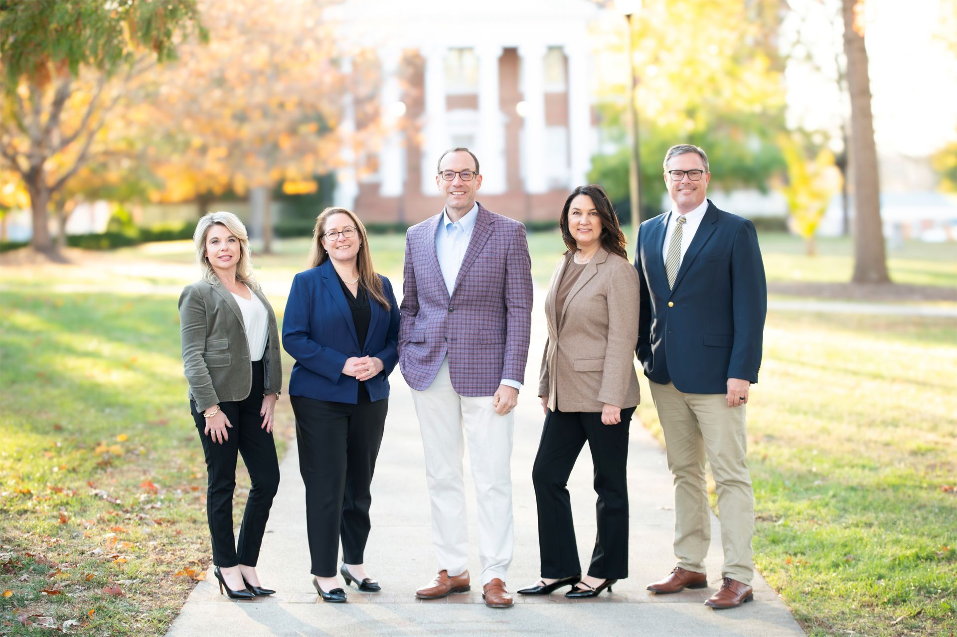 Five people, professionally dressed, stand outside in front of a bush and a brick building.