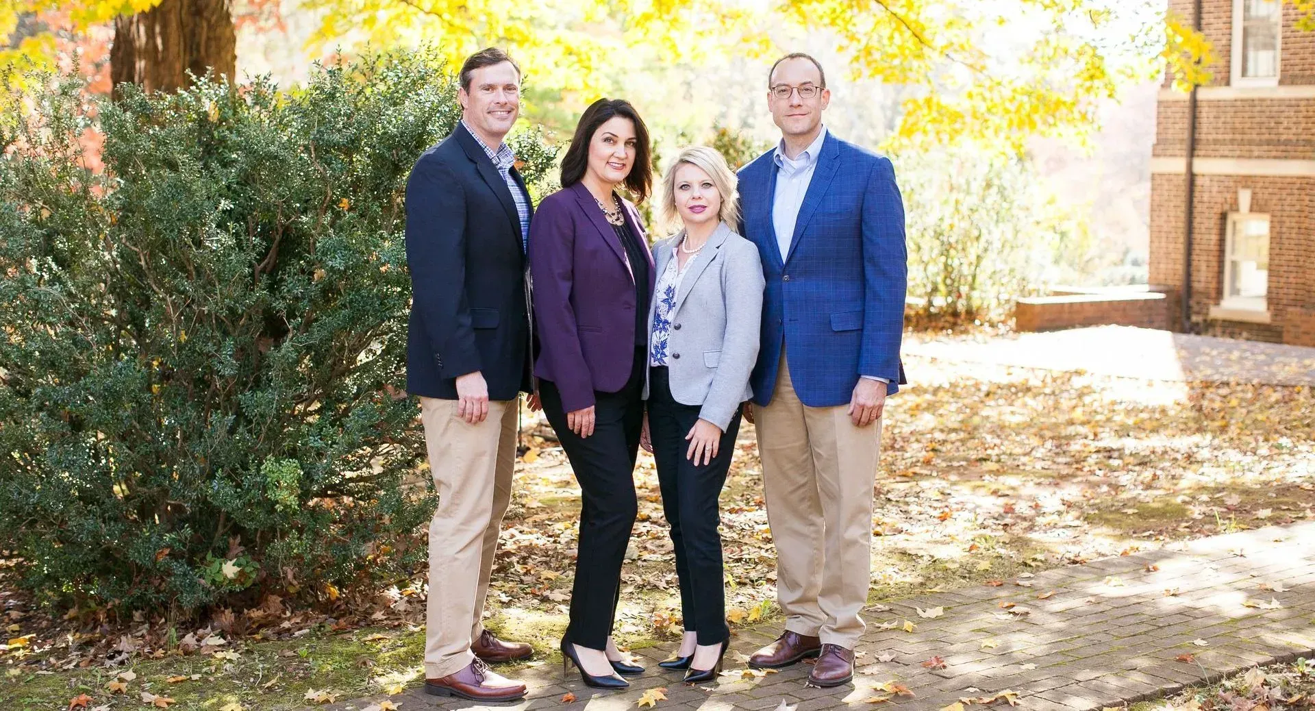 Four people, professionally dressed, stand outside in front of a bush and a brick building.