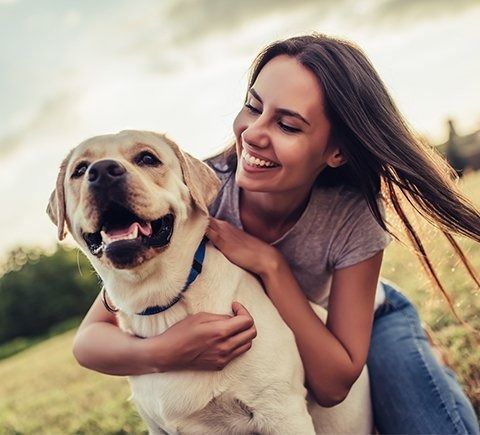 Woman smiling, embracing a yellow Labrador dog in an outdoor setting.