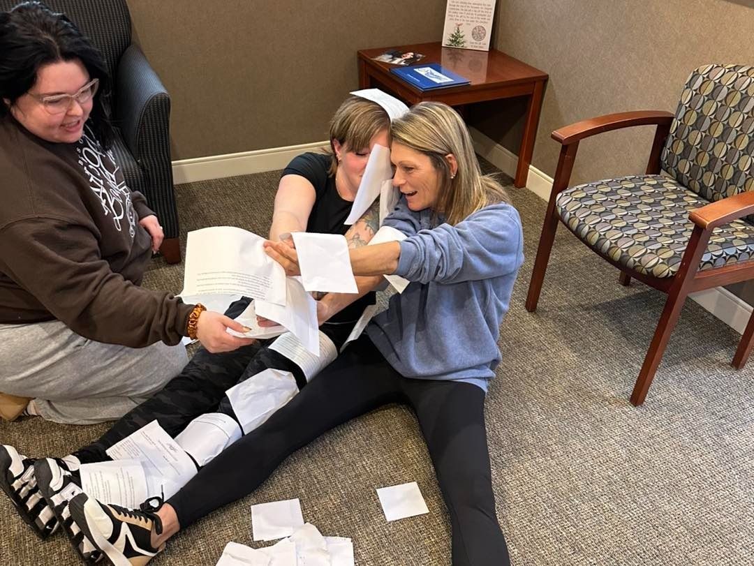 Three people on floor, tearing paper. One holds arms out, another helps. Brown couch, wood chair in room.