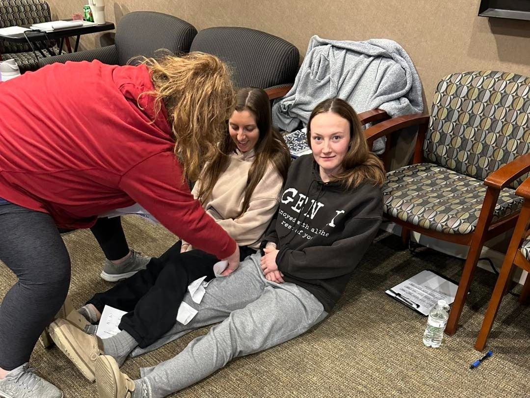 Woman tends to two seated girls on the floor; tan/gray chairs, beige wall; one girl wears gray pants, other, black.