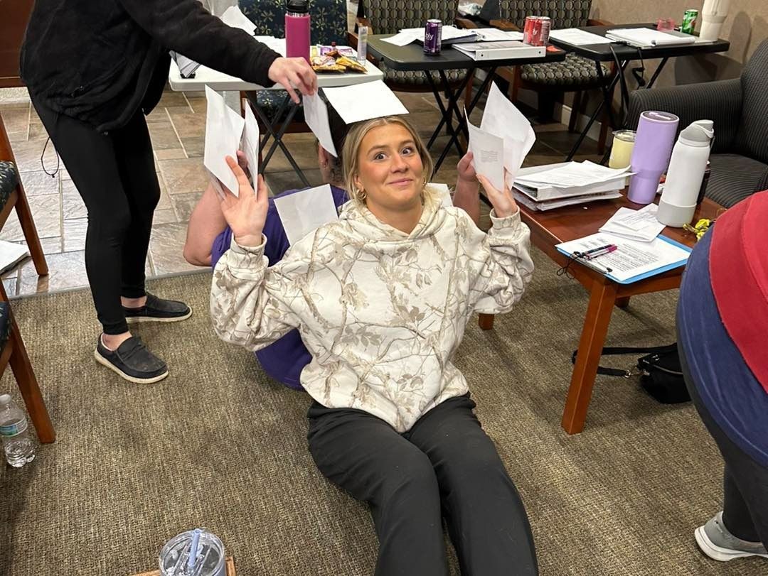 Woman sitting on floor with arms raised, holding papers, smiling. Two other people are in background.
