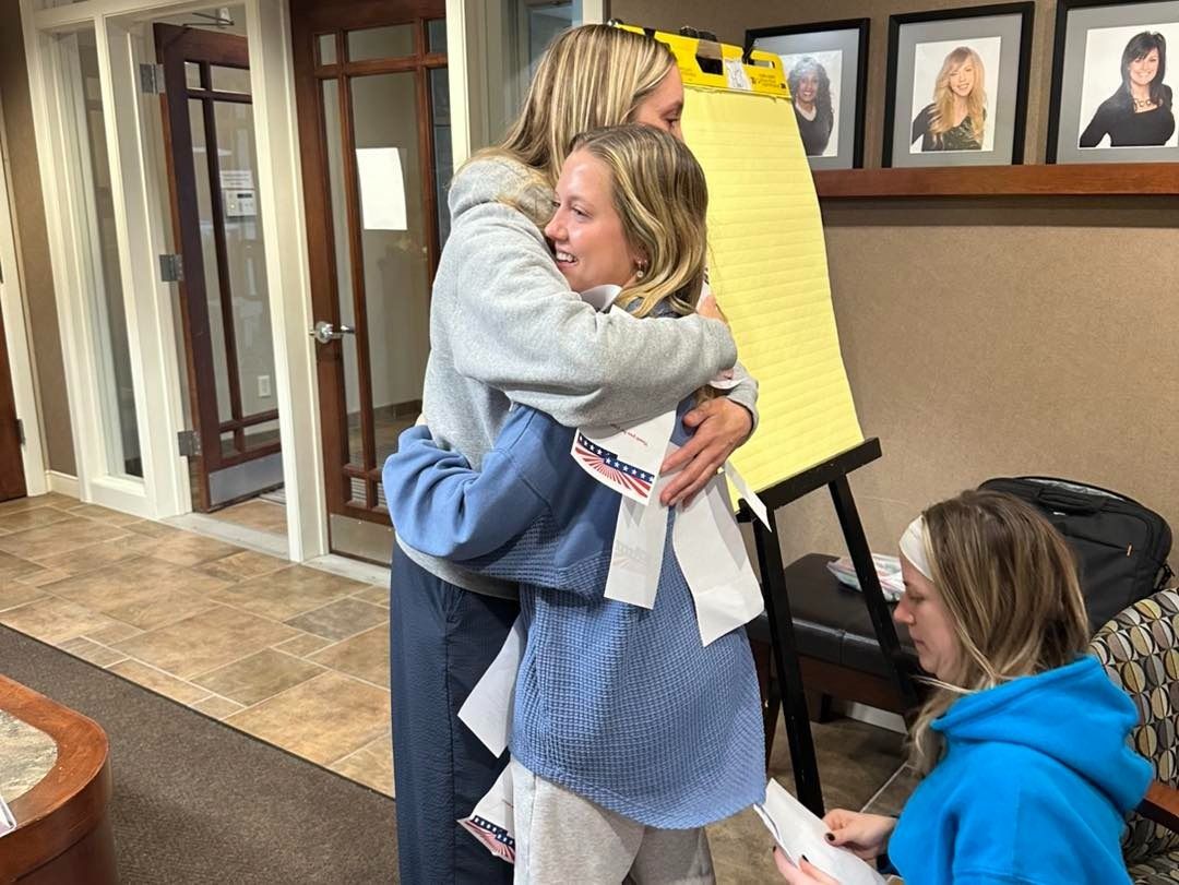 Two women hugging, possibly comforting each other, near a yellow easel and seated woman.