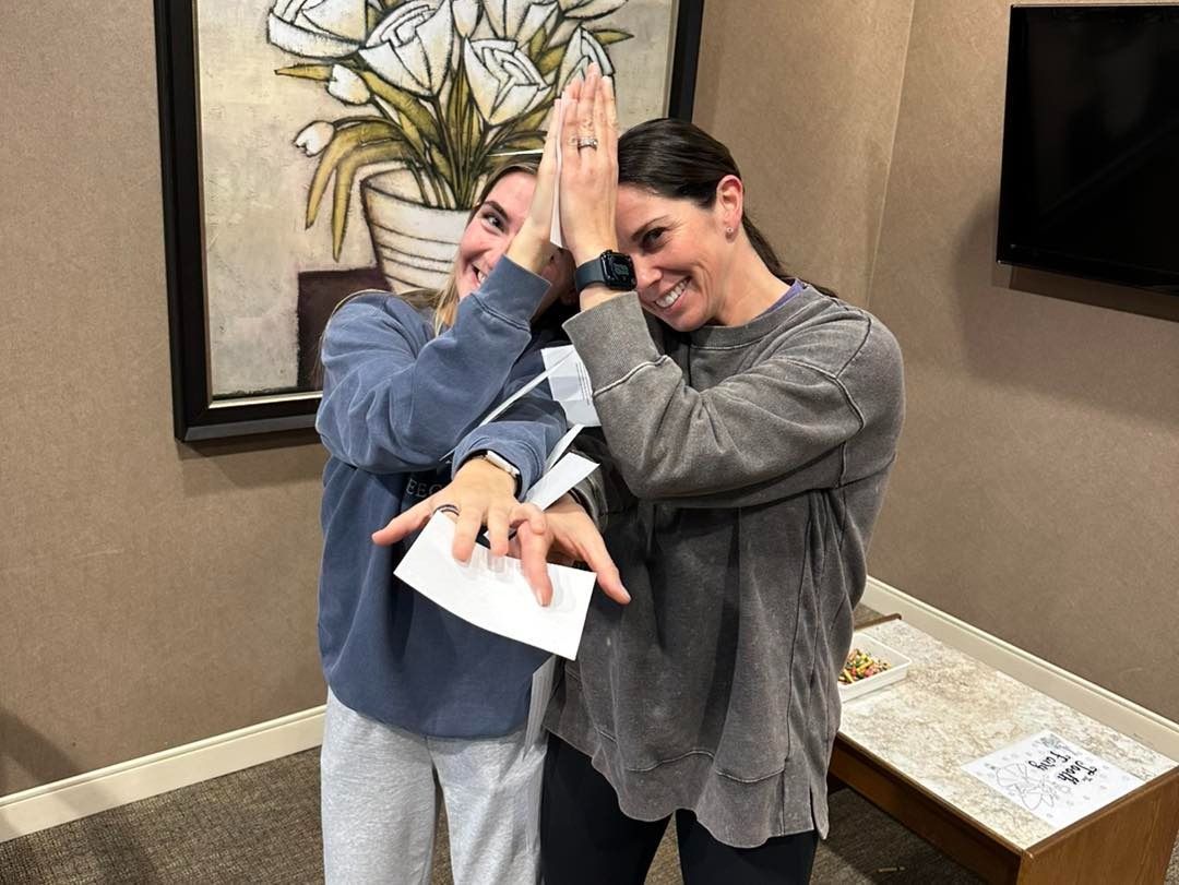 Two women high-fiving, holding paper, smiling. Neutral setting with art and small table.