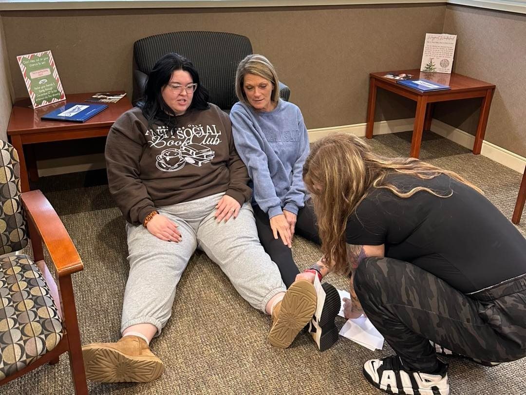 Two women and a person kneel to examine a woman's leg in a room.