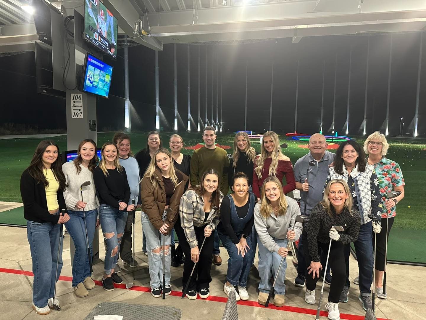 Group of people smiling, posing with golf clubs at an illuminated driving range.