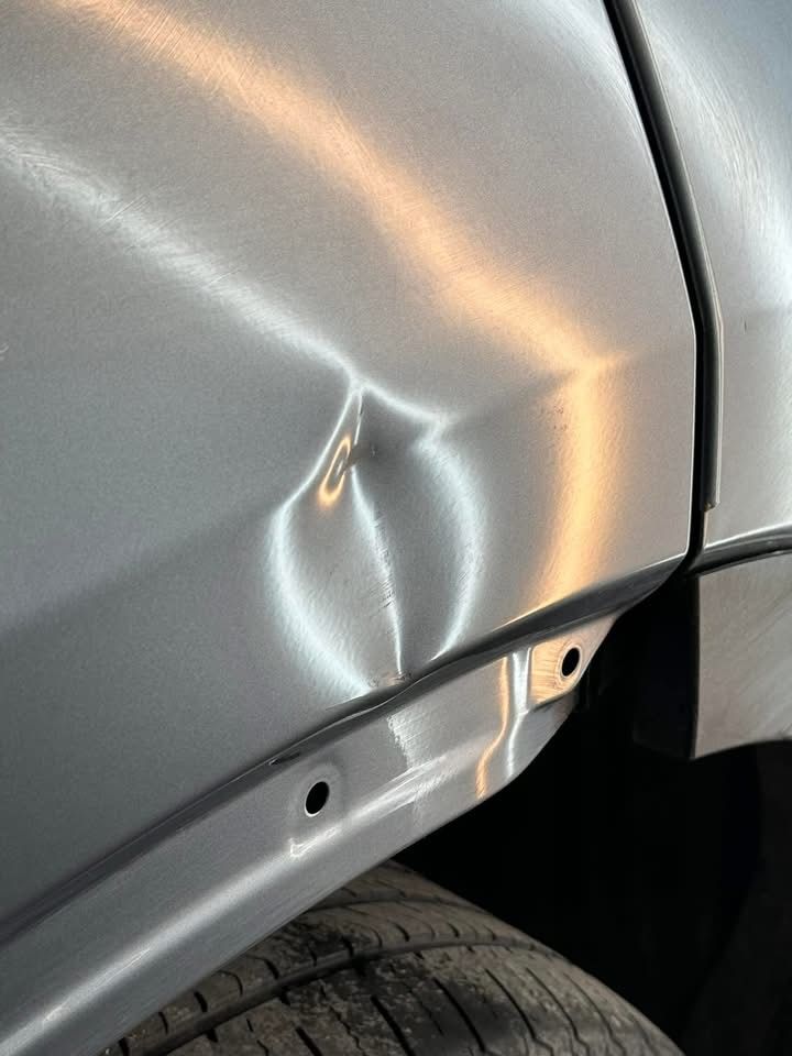 A close up of a silver car with a dent in the fender.