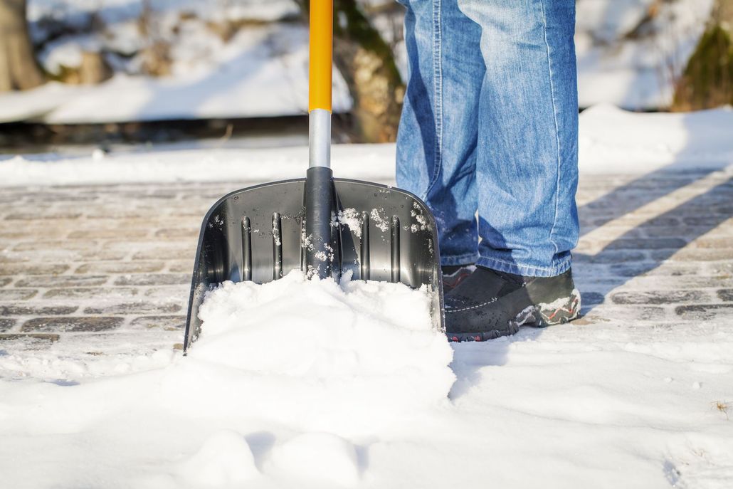 Person shoveling snow from a brick pathway with a black shovel, wearing blue jeans and black boots.