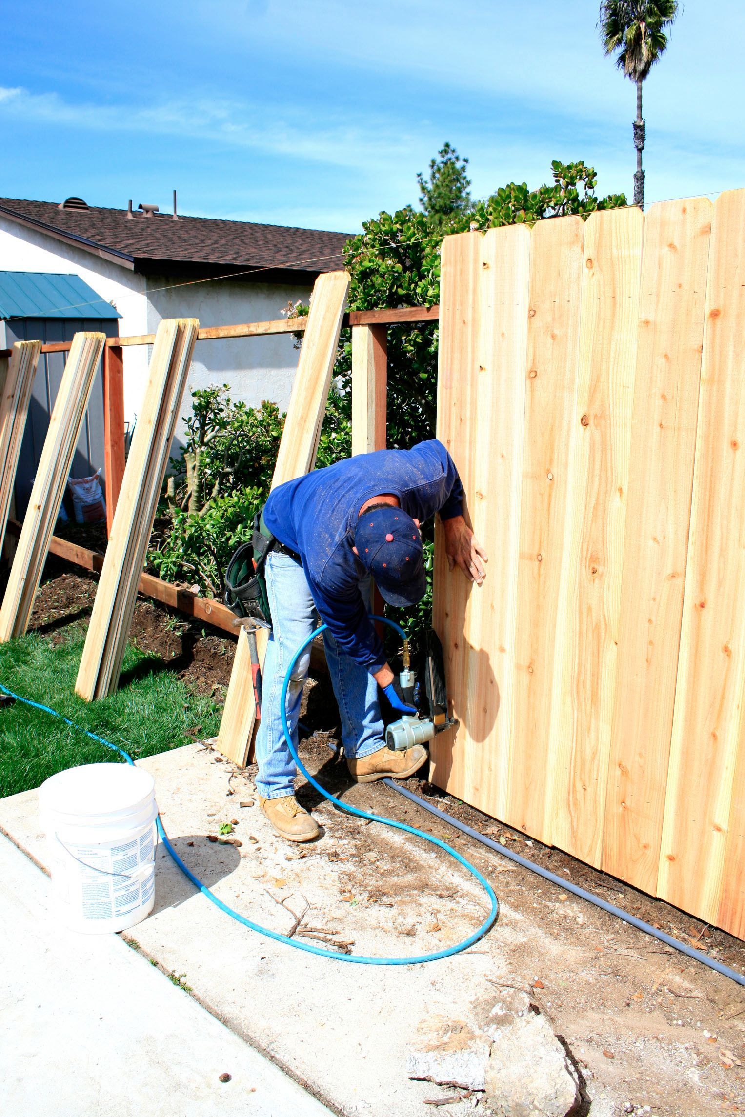 Man building a wooden fence outdoors, using a power tool on a sunny day.