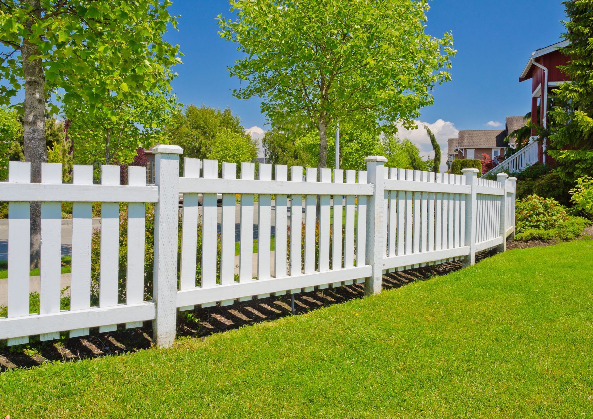 White picket fence on green lawn, trees and houses in the background on a sunny day.
