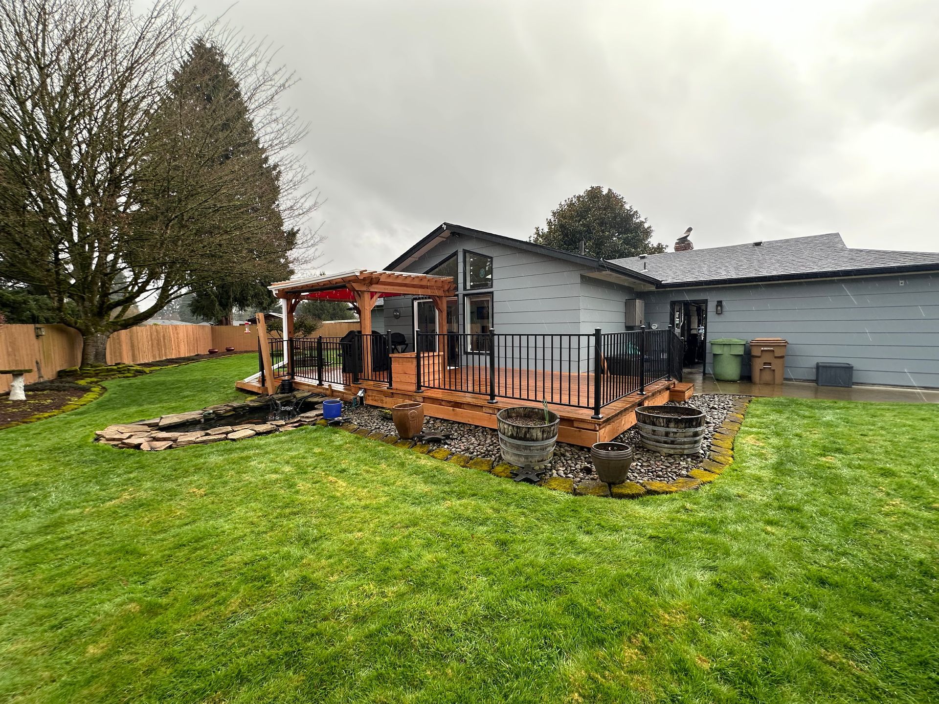 Backyard with a wooden deck, black railing, and a small pond, adjacent to a gray house, on a cloudy day.