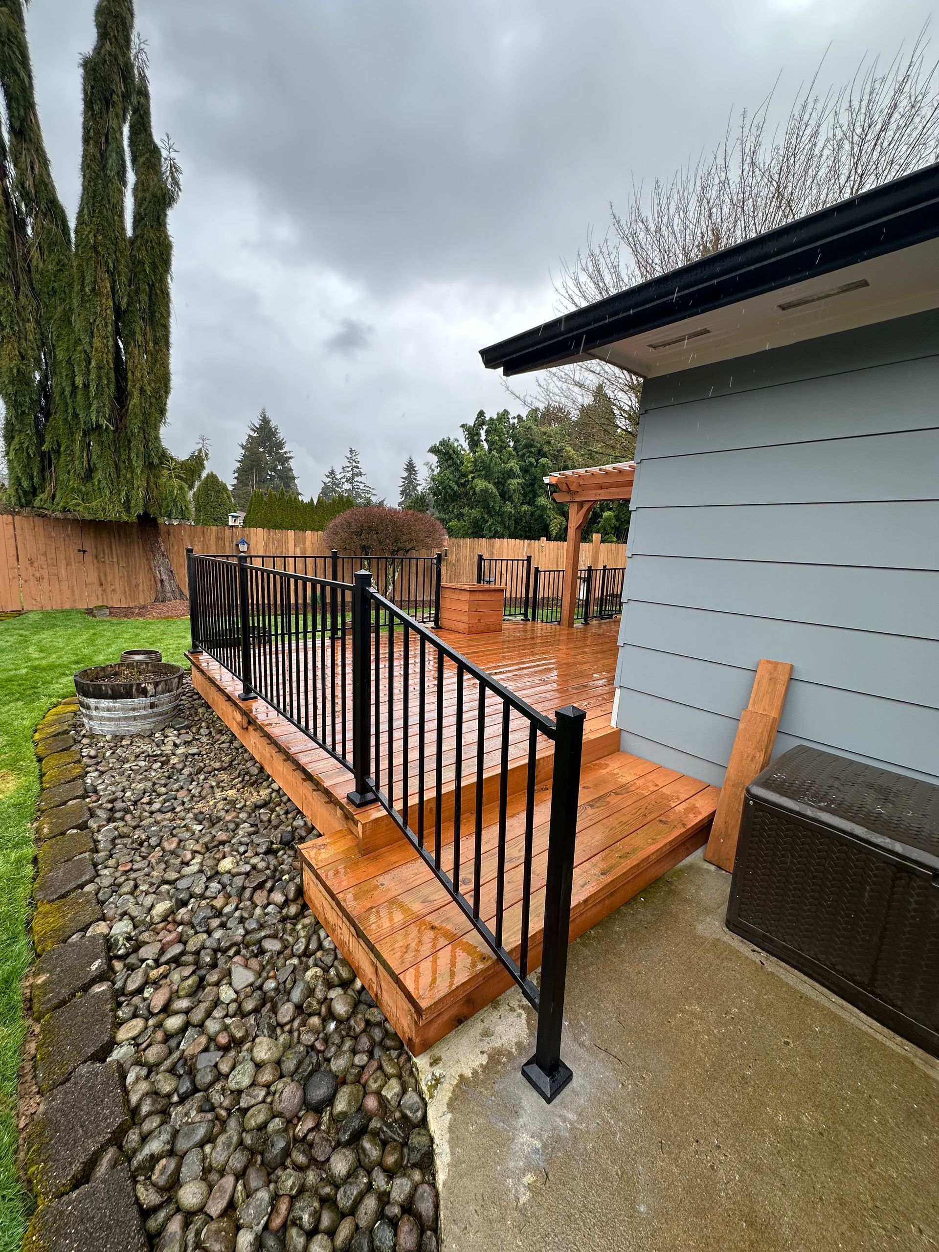 Wooden deck with black railing next to a light blue building and rocky ground, on a cloudy day.