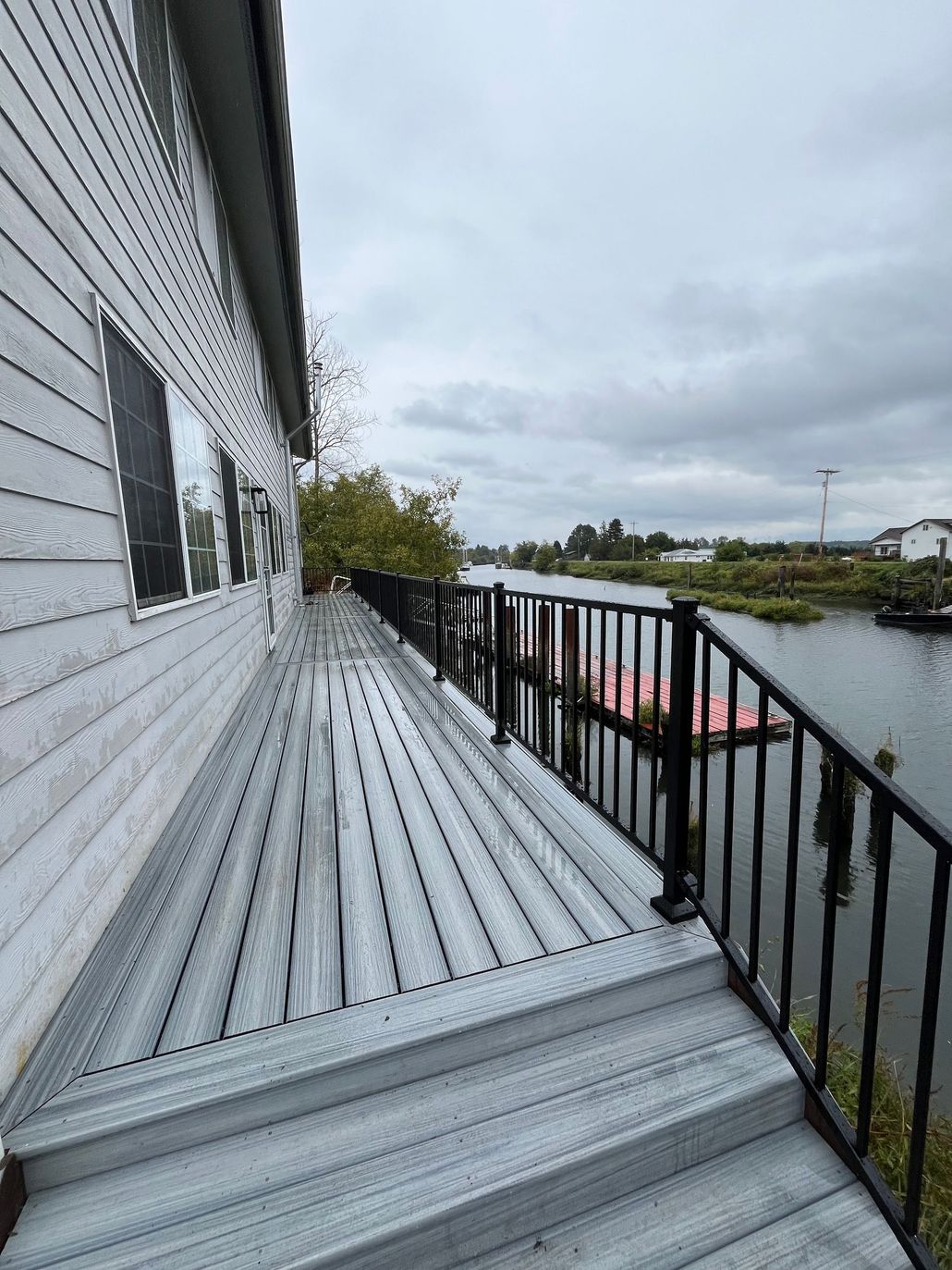 Side of a house with a gray deck overlooking a waterway, with black railing and overcast sky.