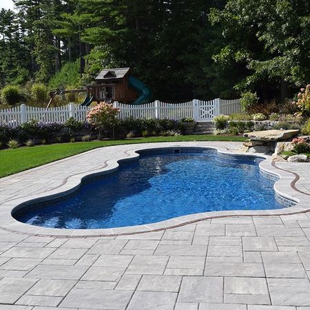 Pool with blue water and stone patio in a backyard with a playground and white picket fence.