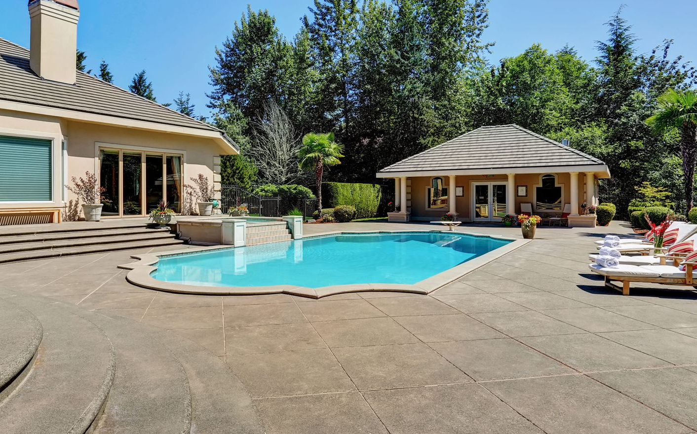 Backyard with a swimming pool, patio, and gazebo, surrounded by trees under a bright blue sky.
