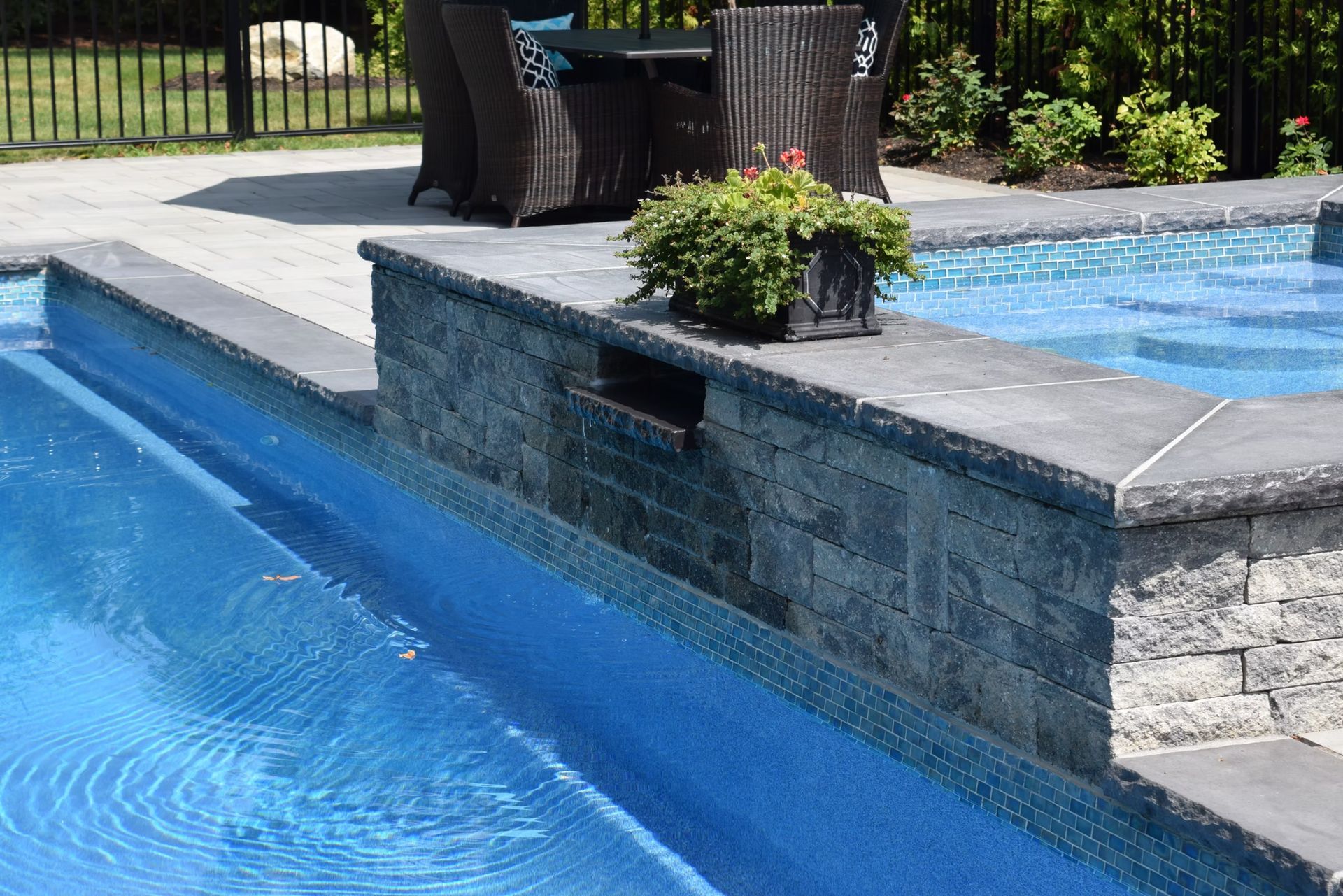 Pool with blue tiles, stone wall with small planter, patio furniture in background.