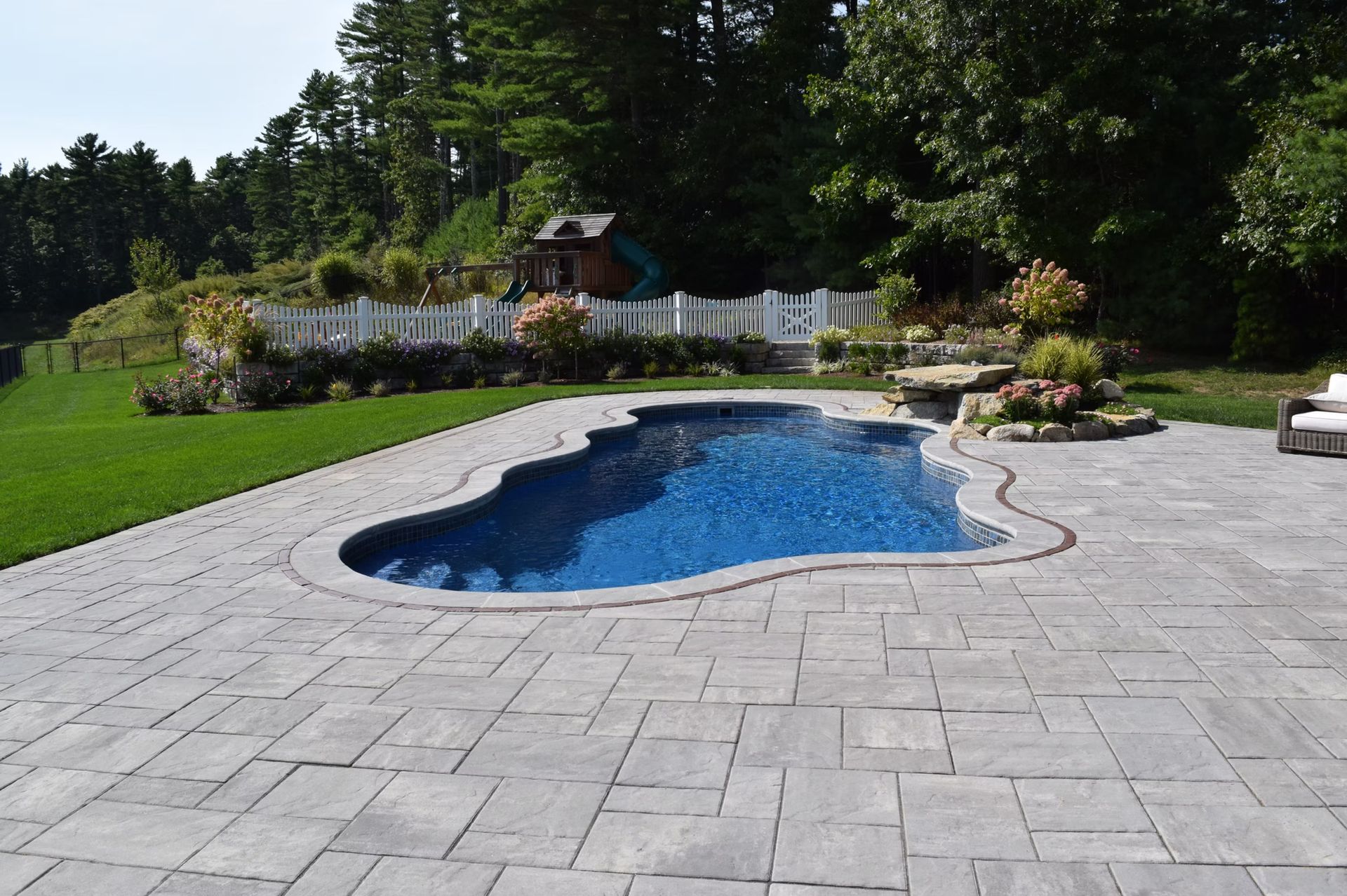 Swimming pool with blue water surrounded by stone patio, green lawn, and trees.