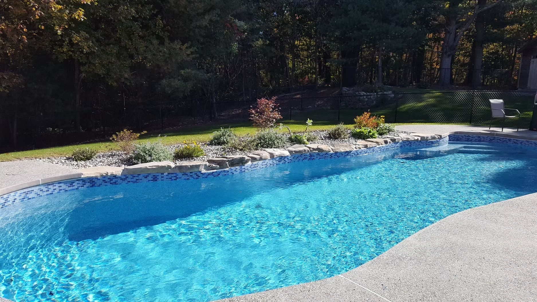 Swimming pool with blue water and stone edging, surrounded by grass, flowers, and trees.