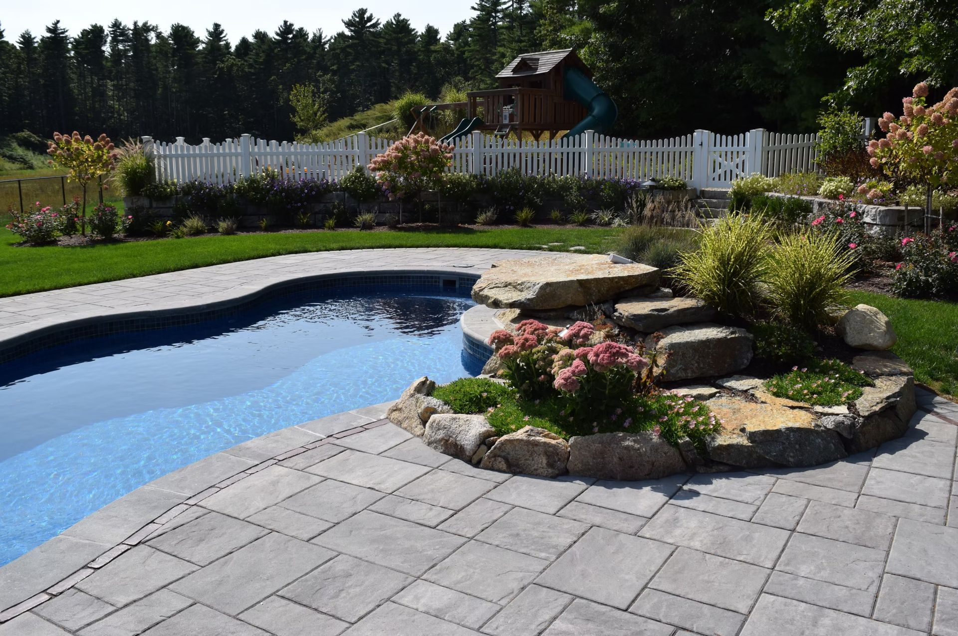 A landscaped backyard with a pool, stone patio, rock steps, and white picket fence.