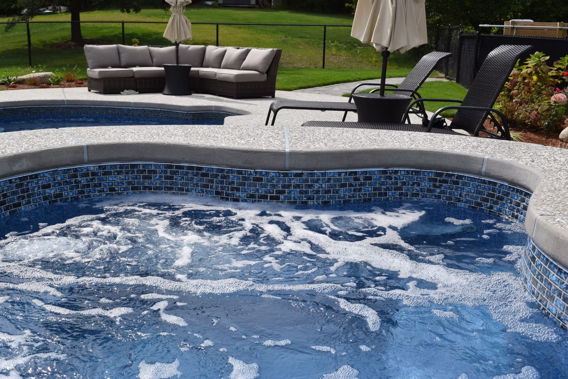 Pool with bubbly water, blue tile, and concrete coping. Wicker outdoor furniture and green grass in the background.