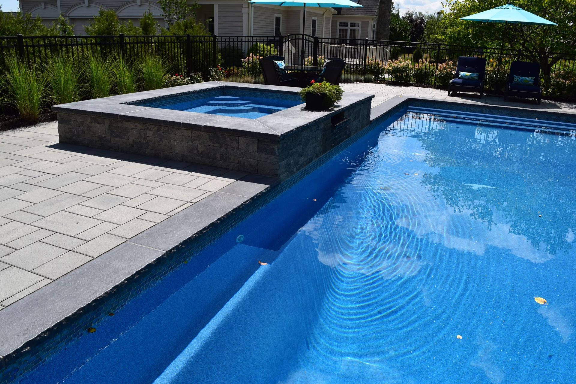 Pool with attached hot tub, blue water, gray stone patio, umbrellas, and landscaping.