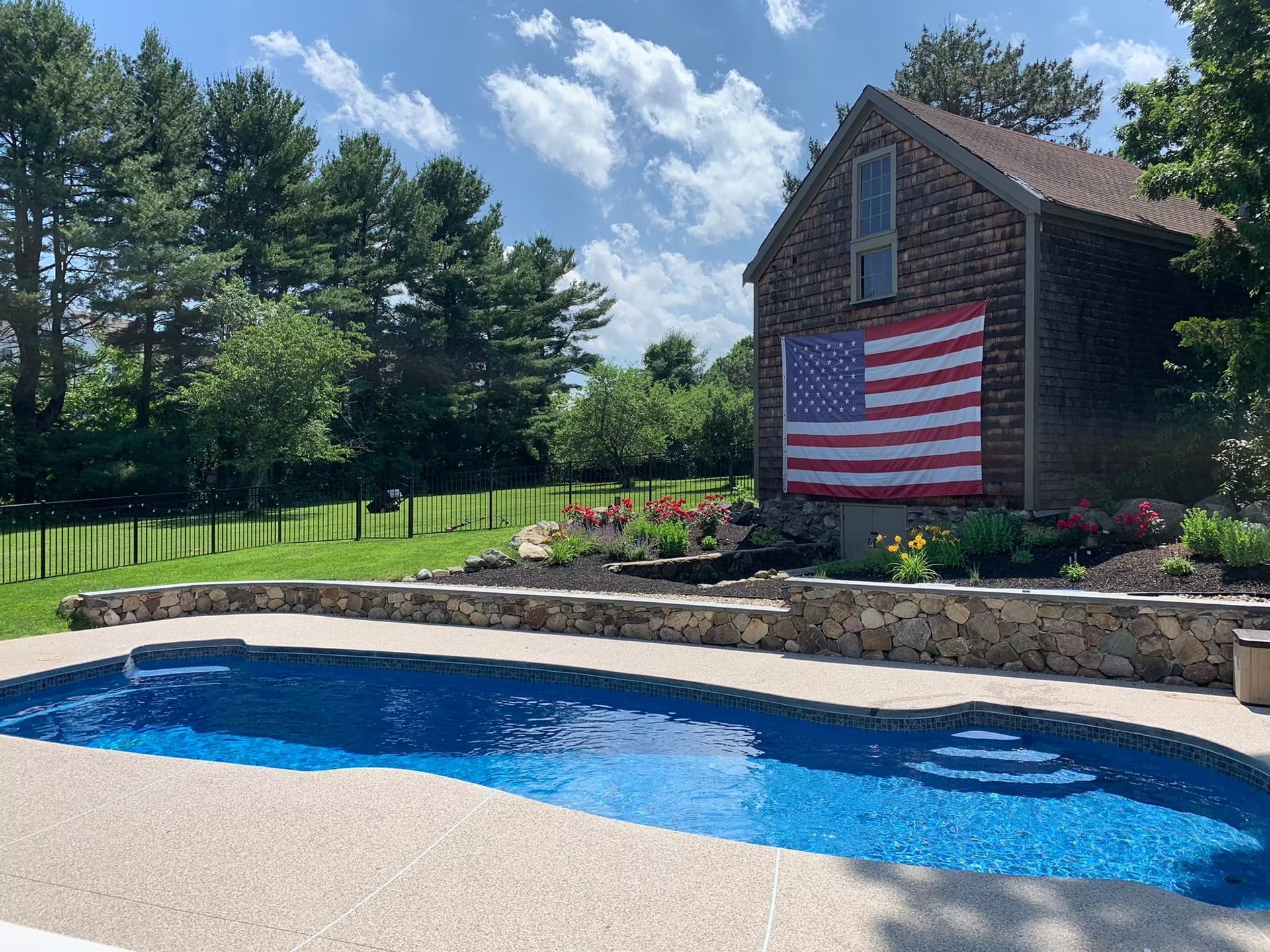 A backyard pool with blue water, an American flag on a barn, and a sunny sky.