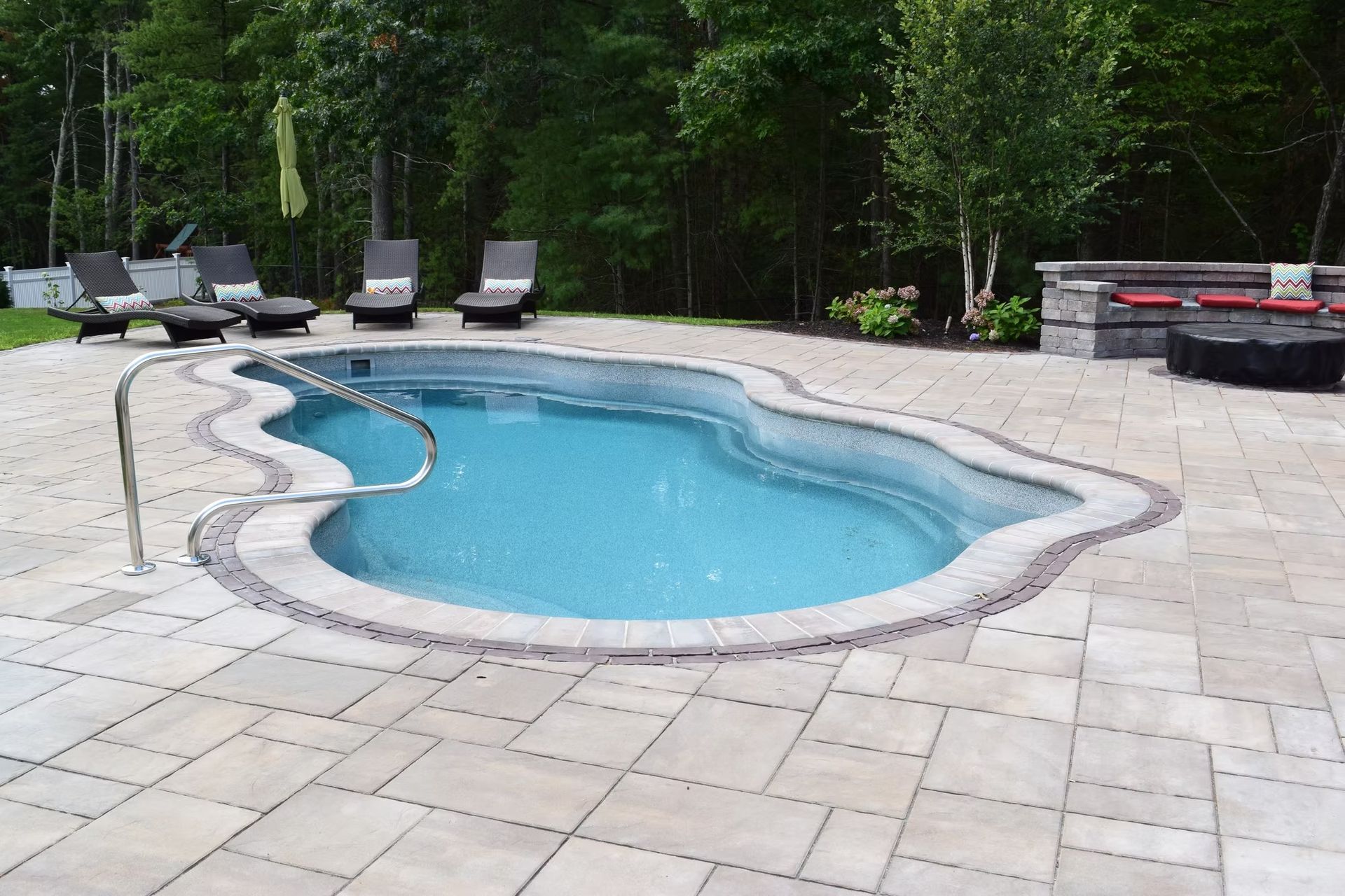 Swimming pool with a curved shape, surrounded by light-colored paving stones. Lounge chairs and a fire pit visible.
