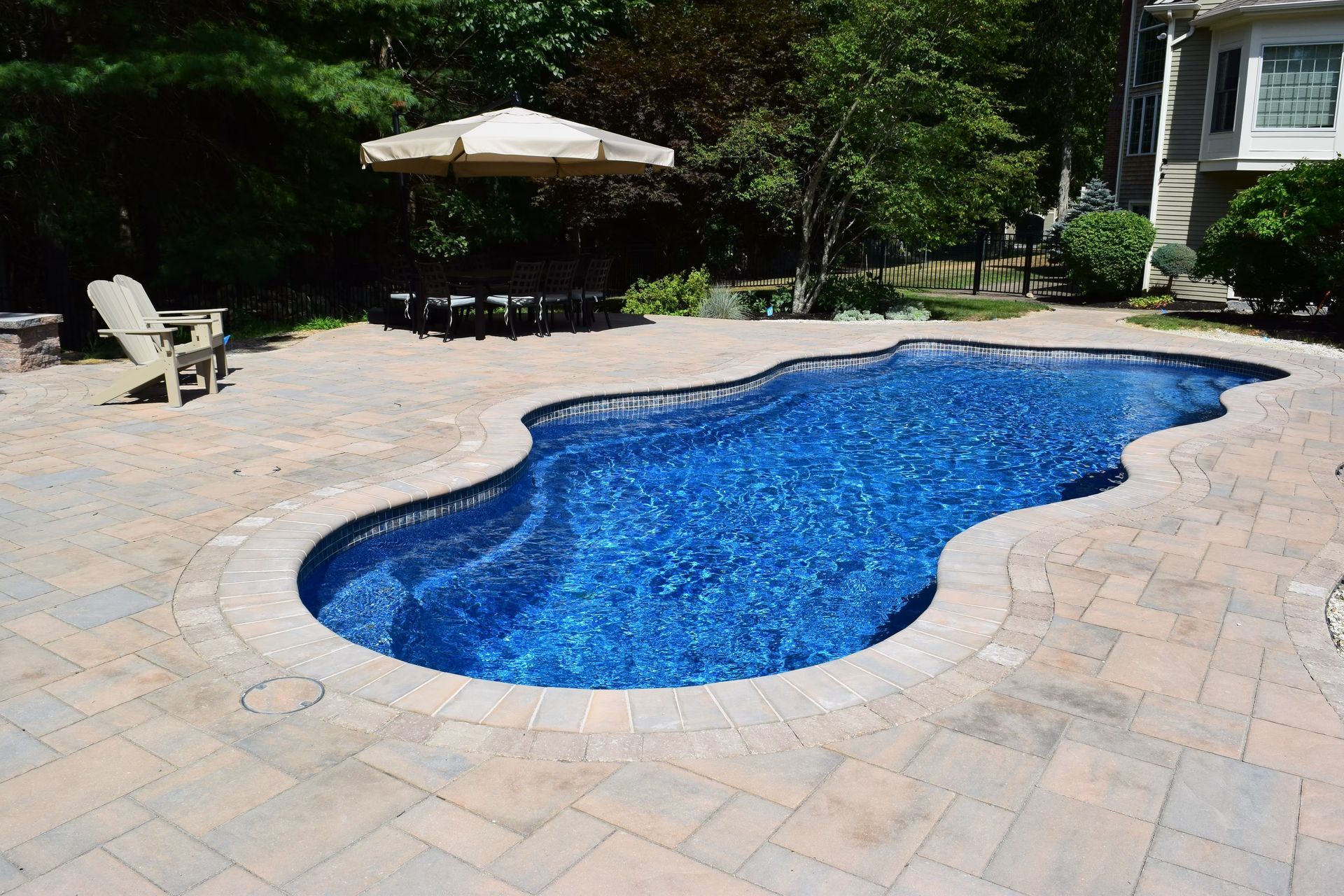 Pool surrounded by stone patio, with umbrella and chair nearby, trees in the background.