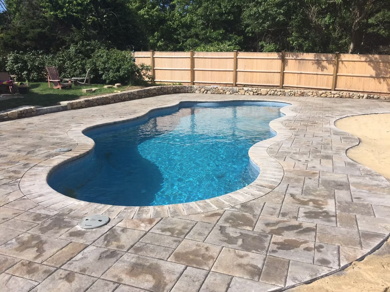 Swimming pool with blue water surrounded by stone pavers, grass, and a wooden fence.