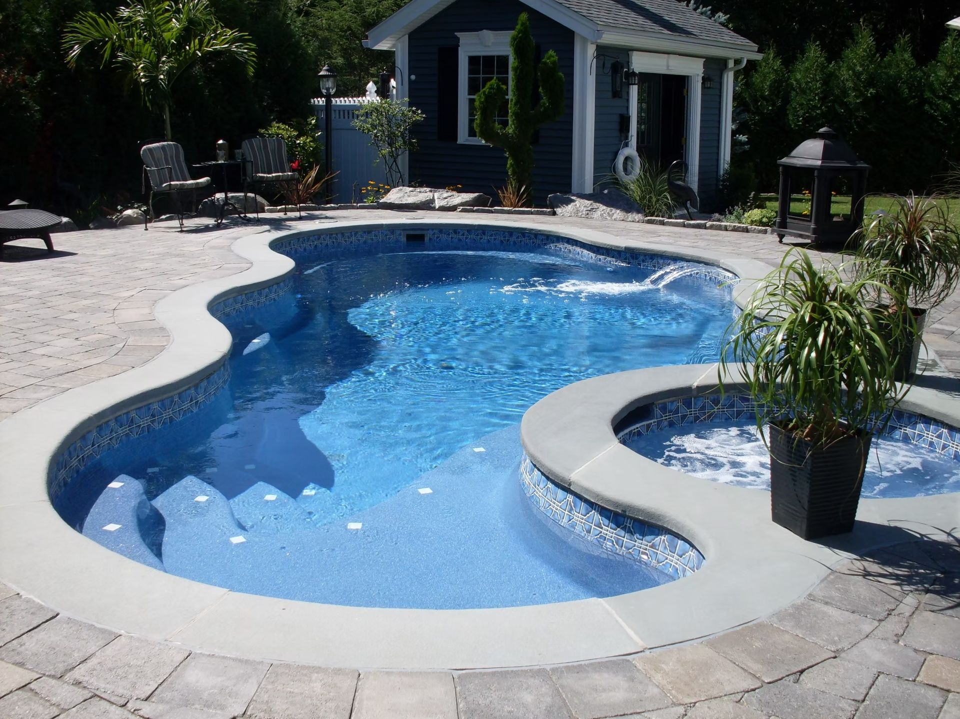 Blue, freeform swimming pool with attached spa and steps, surrounded by stone patio, small blue shed in the background.