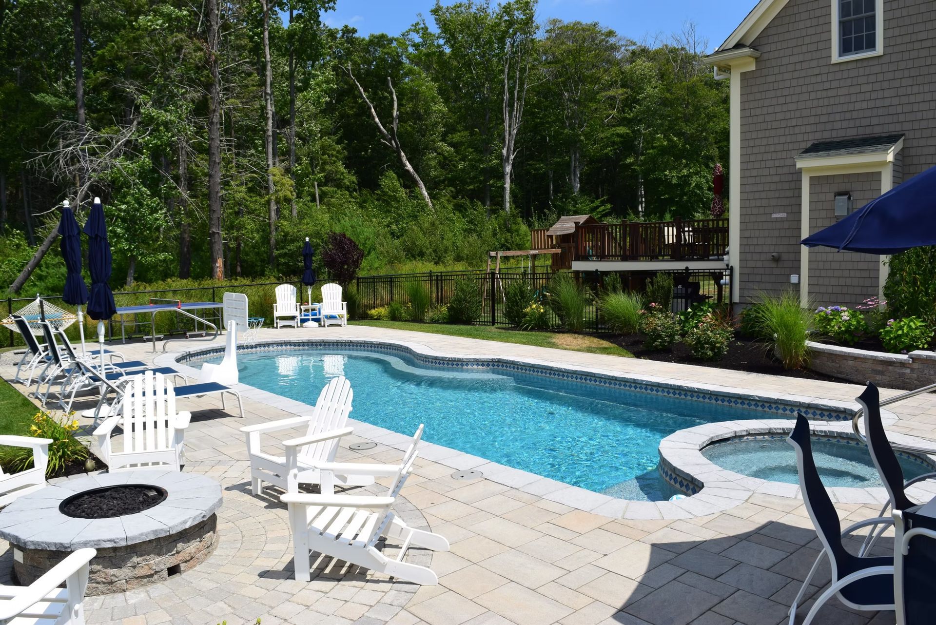 Backyard pool with patio, fire pit, and white Adirondack chairs; lush green trees in the background.