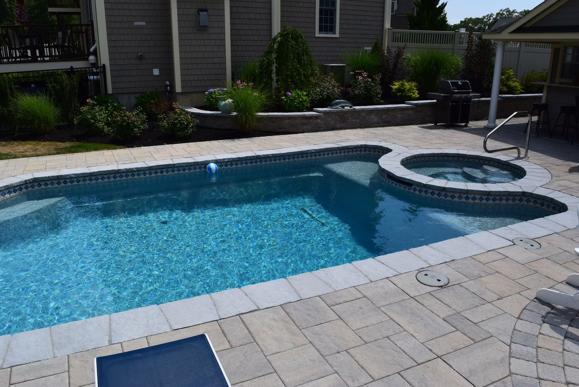 Swimming pool with attached hot tub, surrounded by stone patio. Backyard setting, blue water.