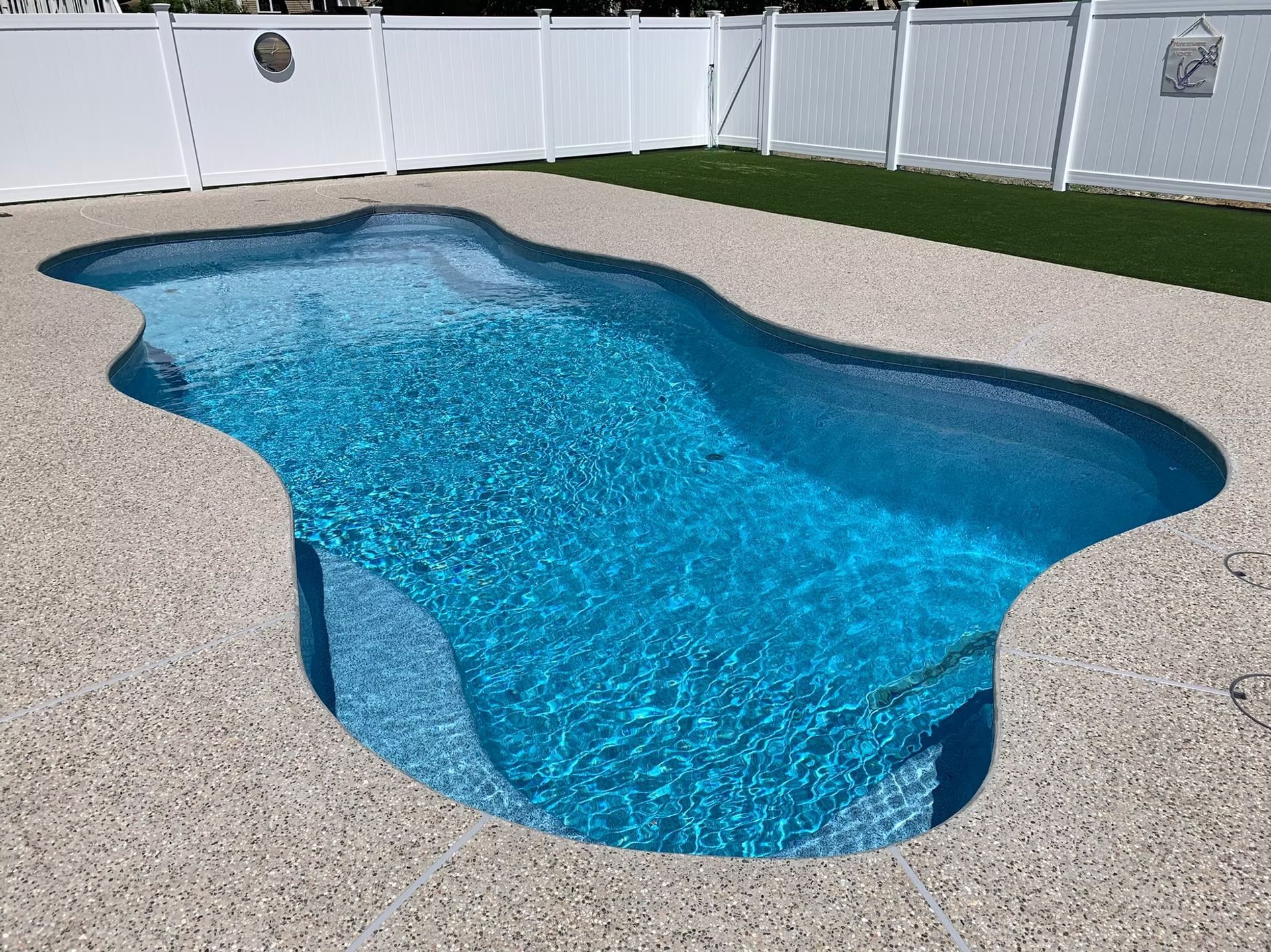 A free-form swimming pool with turquoise water, surrounded by tan concrete and a white fence.