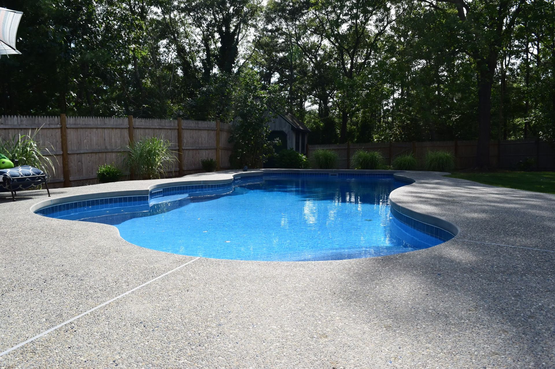 Swimming pool with blue water surrounded by a concrete patio and a wooden fence.