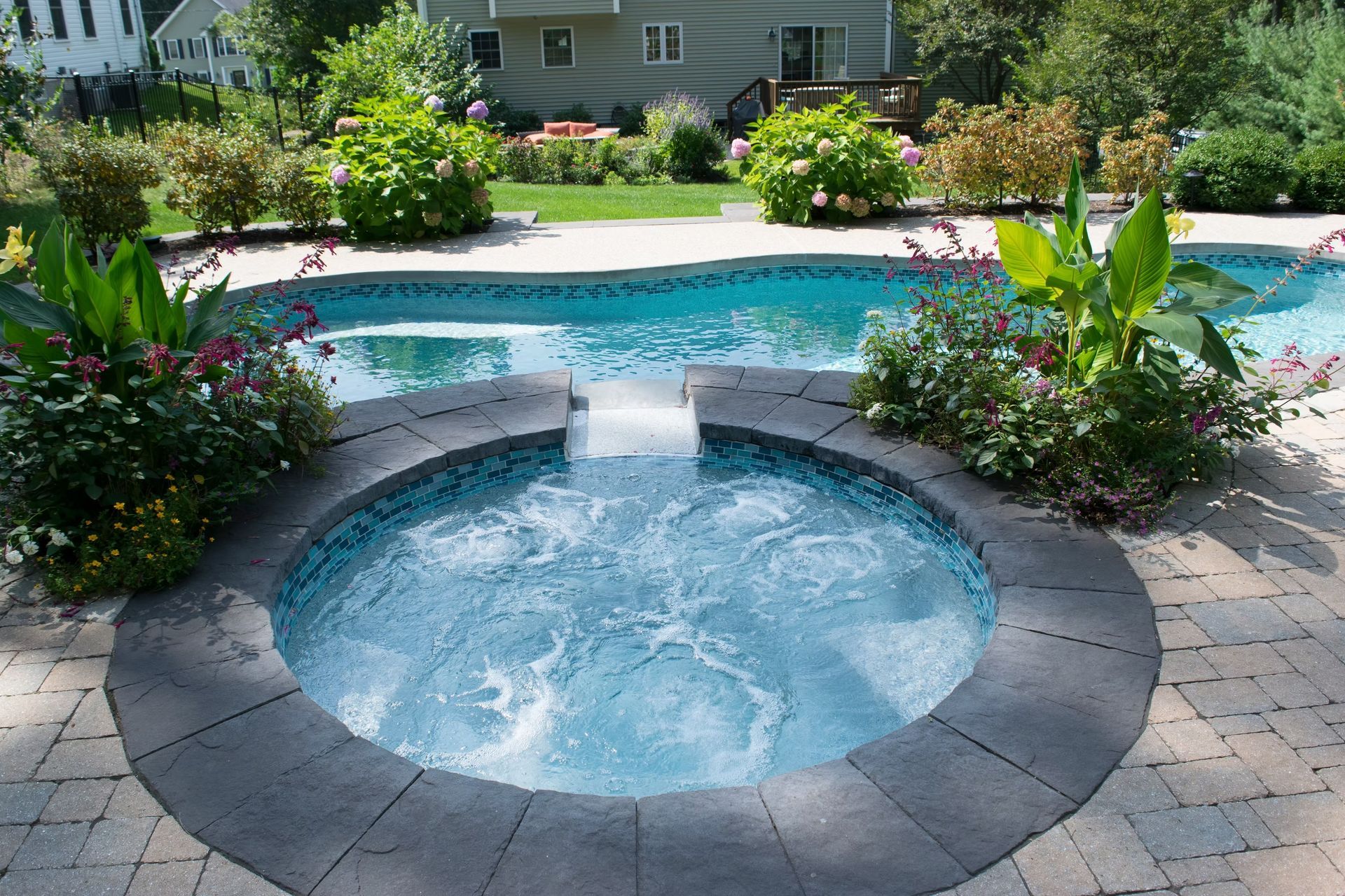 Pool with a connected spa, surrounded by stone and plants, backyard setting.