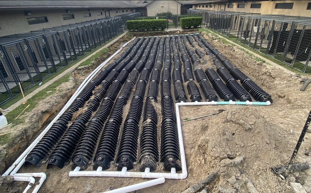 Installation of black, cylindrical septic system components in a dirt trench, surrounded by buildings and landscaping.