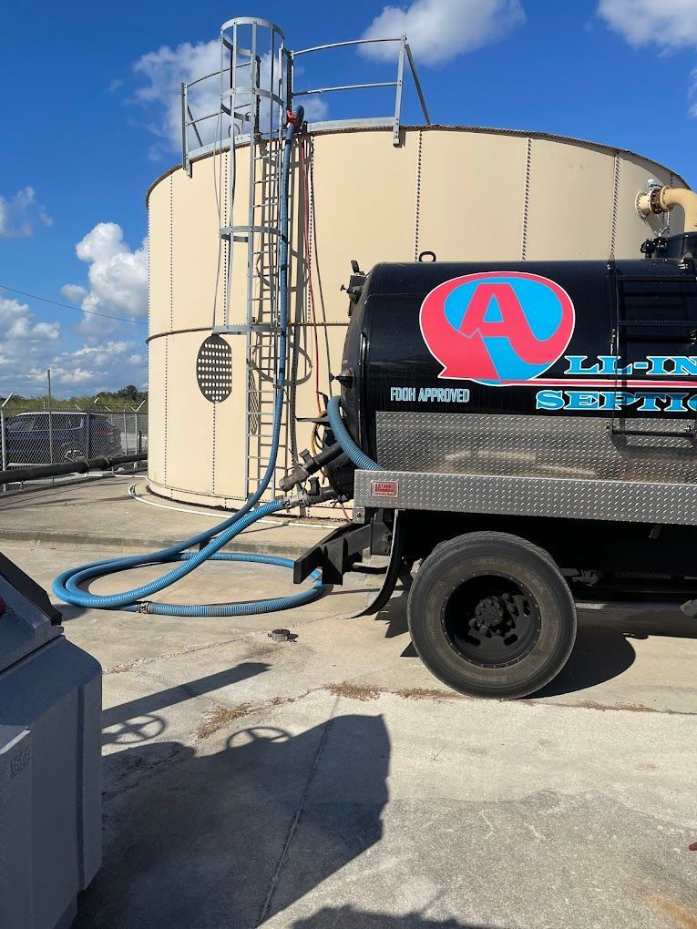 Black tanker truck pumping fluid from beige tank; blue hose connected. Sunny outdoor setting.