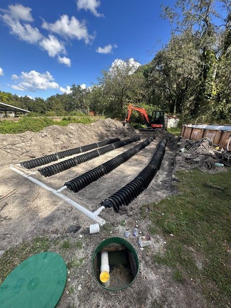 Construction site with black drainage pipes laid in trenches, an excavator, and a septic tank lid.