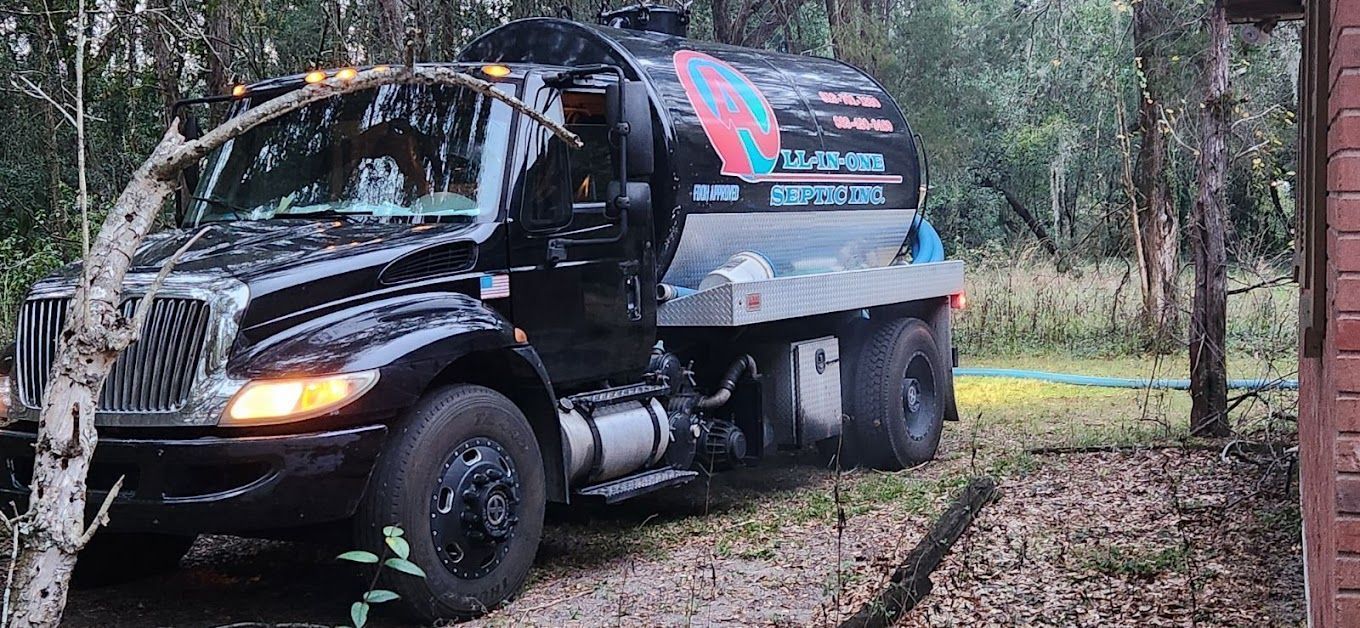 Black tanker truck parked on grassy ground, partially obscured by trees.