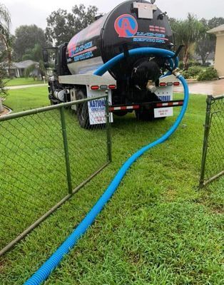A septic tank pumping truck with a blue hose on green grass next to a chain-link fence.