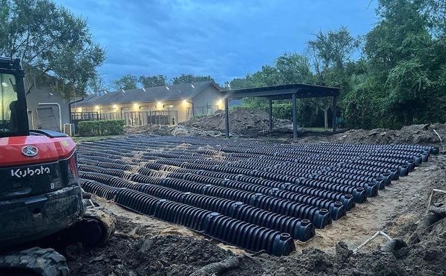 Rows of black drainage pipes laid out in a dirt field with a small excavator, building and trees in the background.