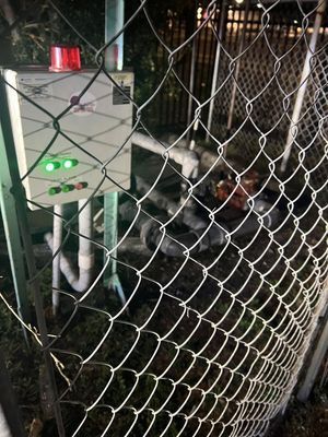 Control panel with flashing red light and green lights, behind a chain link fence.