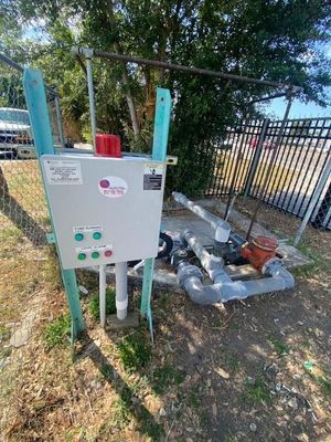 Outdoor water pump control panel with plumbing, supported by teal metal framework.