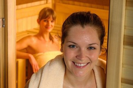 A woman is smiling in a sauna while another woman is wrapped in a towel.