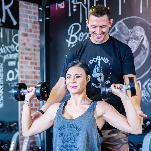 Woman lifting dumbbells with trainer's guidance in a boxing gym. Both smiling.