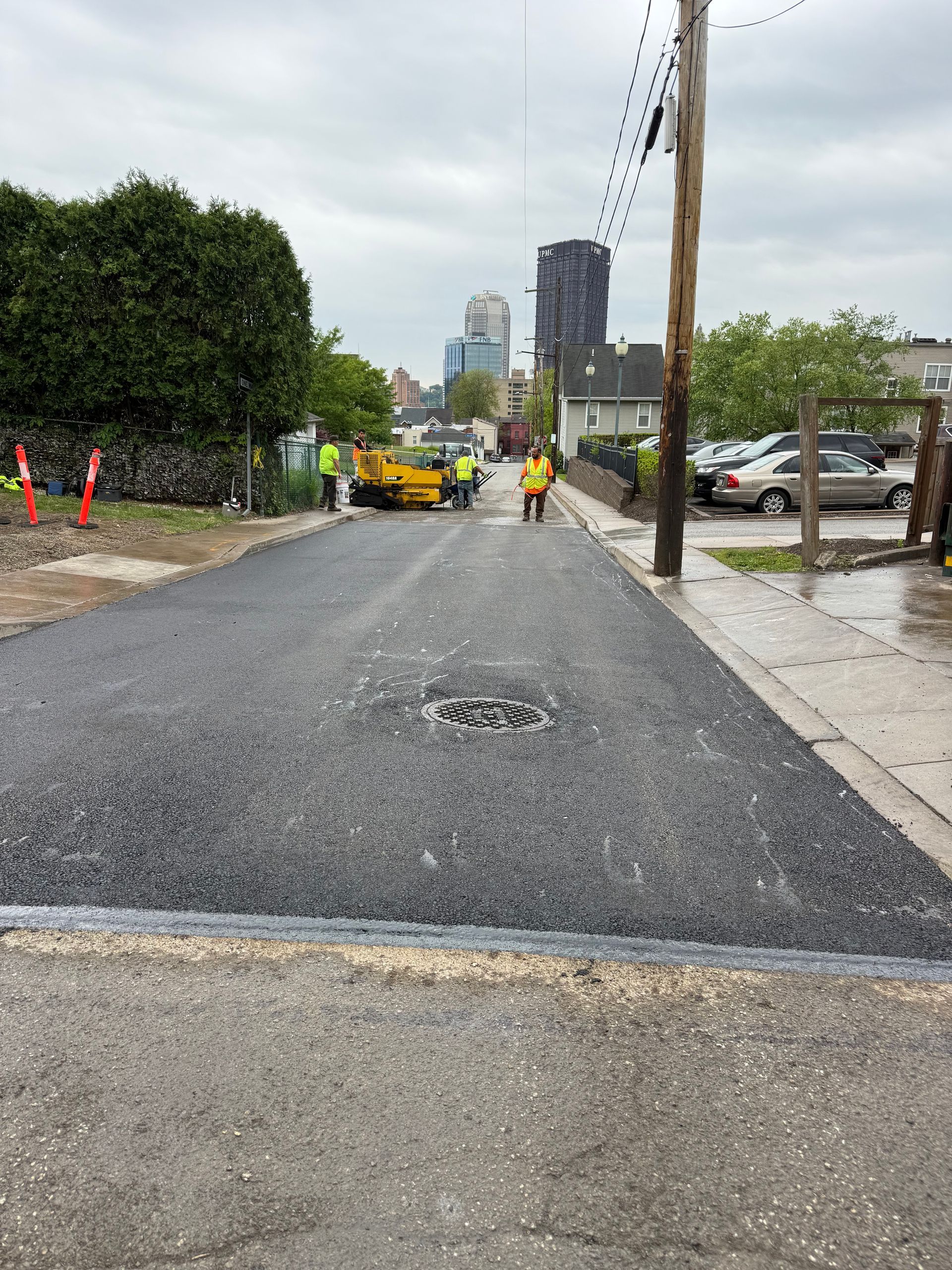 Roadwork on a street with workers, a yellow truck, and fresh asphalt under an overcast sky