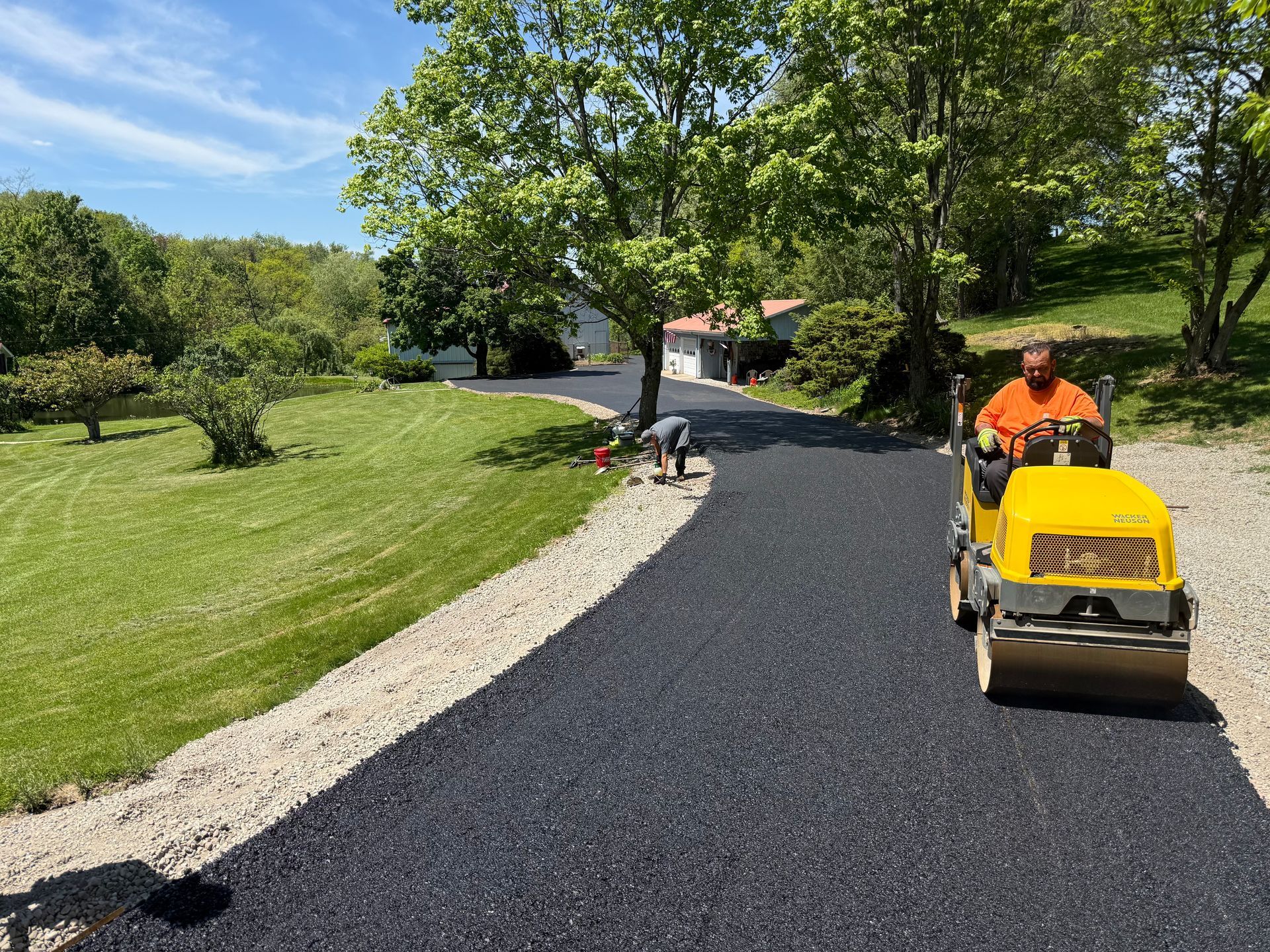 Worker compacting fresh black driveway beside a green lawn and trees