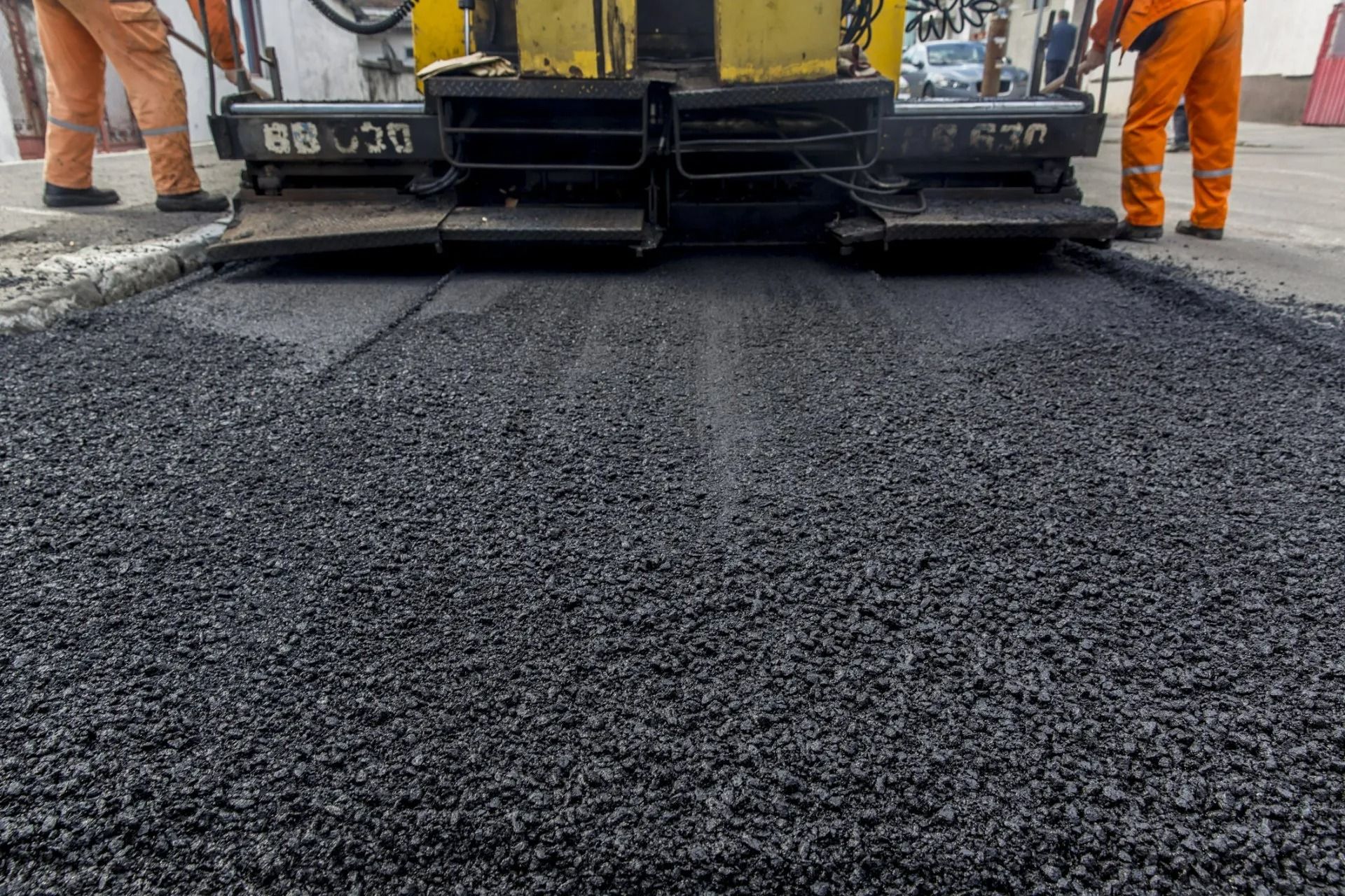 Asphalt paving machine laying fresh asphalt on a road; workers in orange clothing nearby.