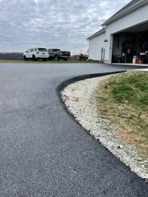 Paved driveway curves toward a white garage. Gravel edge next to green grass. Two trucks are parked nearby.